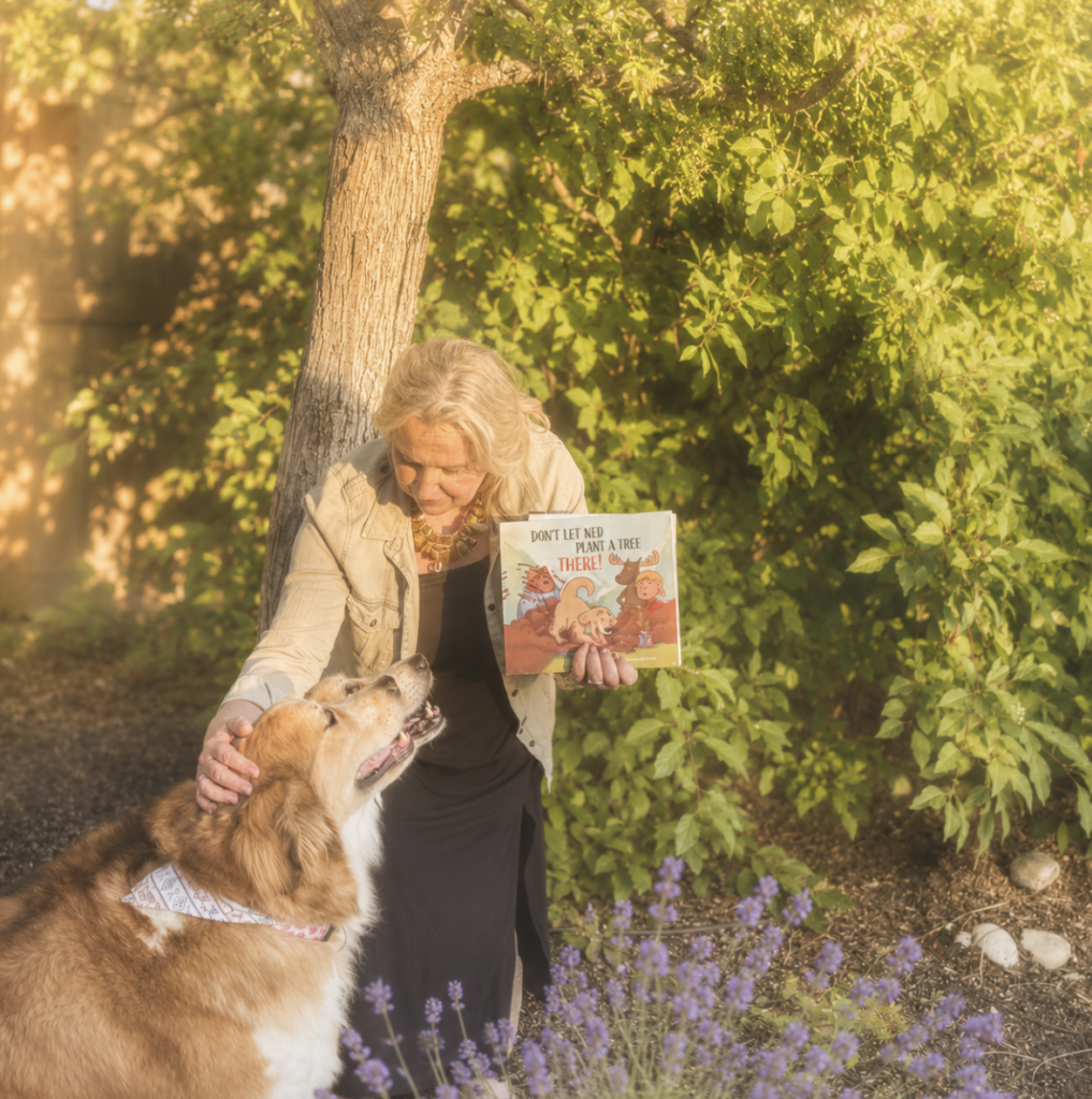 Author Andrea Koehle Jones bends over to greet her two maountain dogs while showing them the book they inspired, Don't Let Ned Plant a Tree THERE! 
